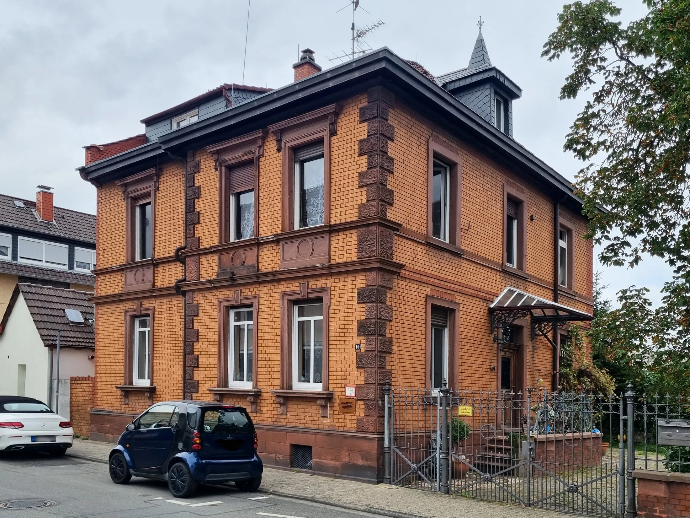 Denkmalgeschütztes zweigeschossiges Wohnhaus aus rotem Backstein in der Pfungstädter Straße in Griesheim, mit Mansarddach, Erker und Gartentor, davor zwei geparkte Autos.