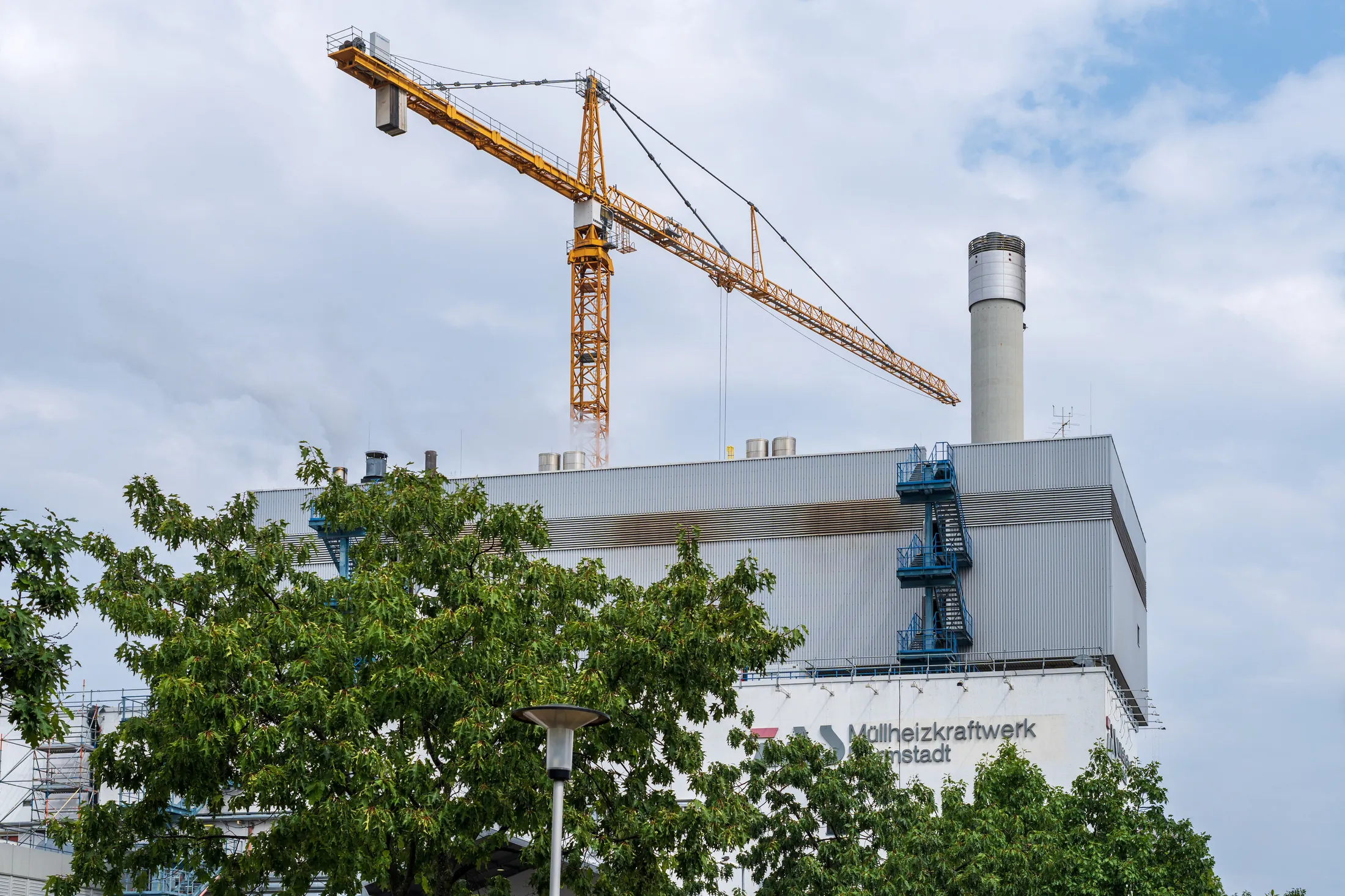 Das Müllheizkraftwerk Darmstadt im August 2026, ein hellgrauer Kraftwerksbau mit Schornstein und Baukran, davor Baumkronen und eine Straßenlaterne.