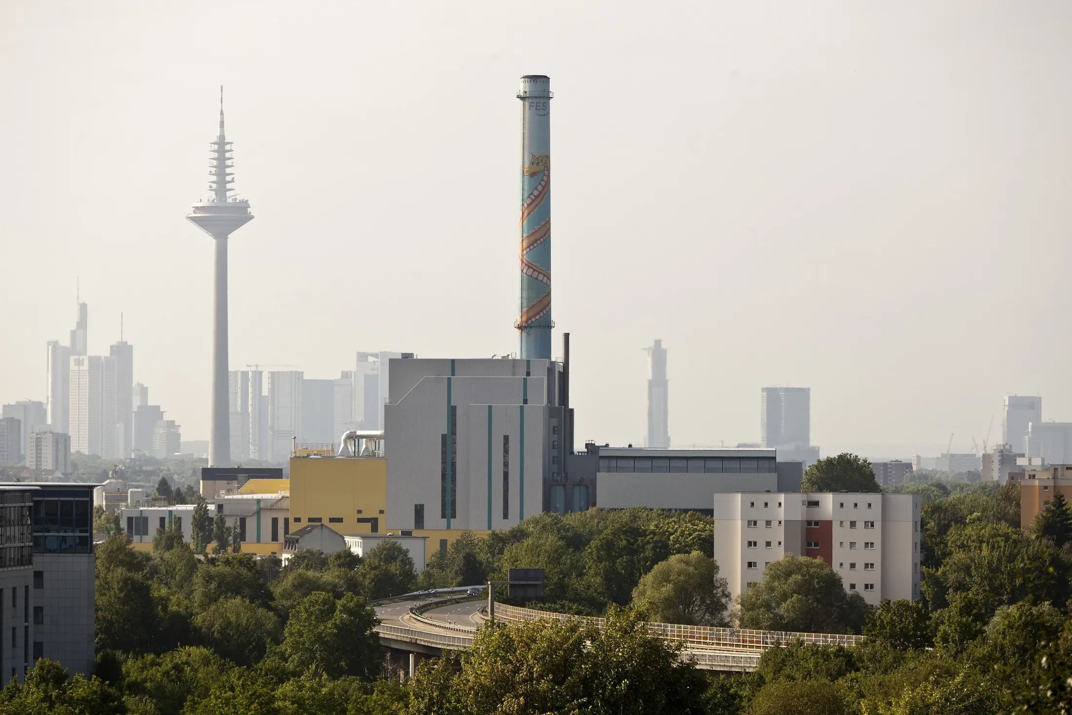Das Müllheizkraftwerk Nordweststadt mit seinem hohen Schornstein, dahinter der Europaturm und die Frankfurter Hochhäuser im Dunst, im Vordergrund Bäume.