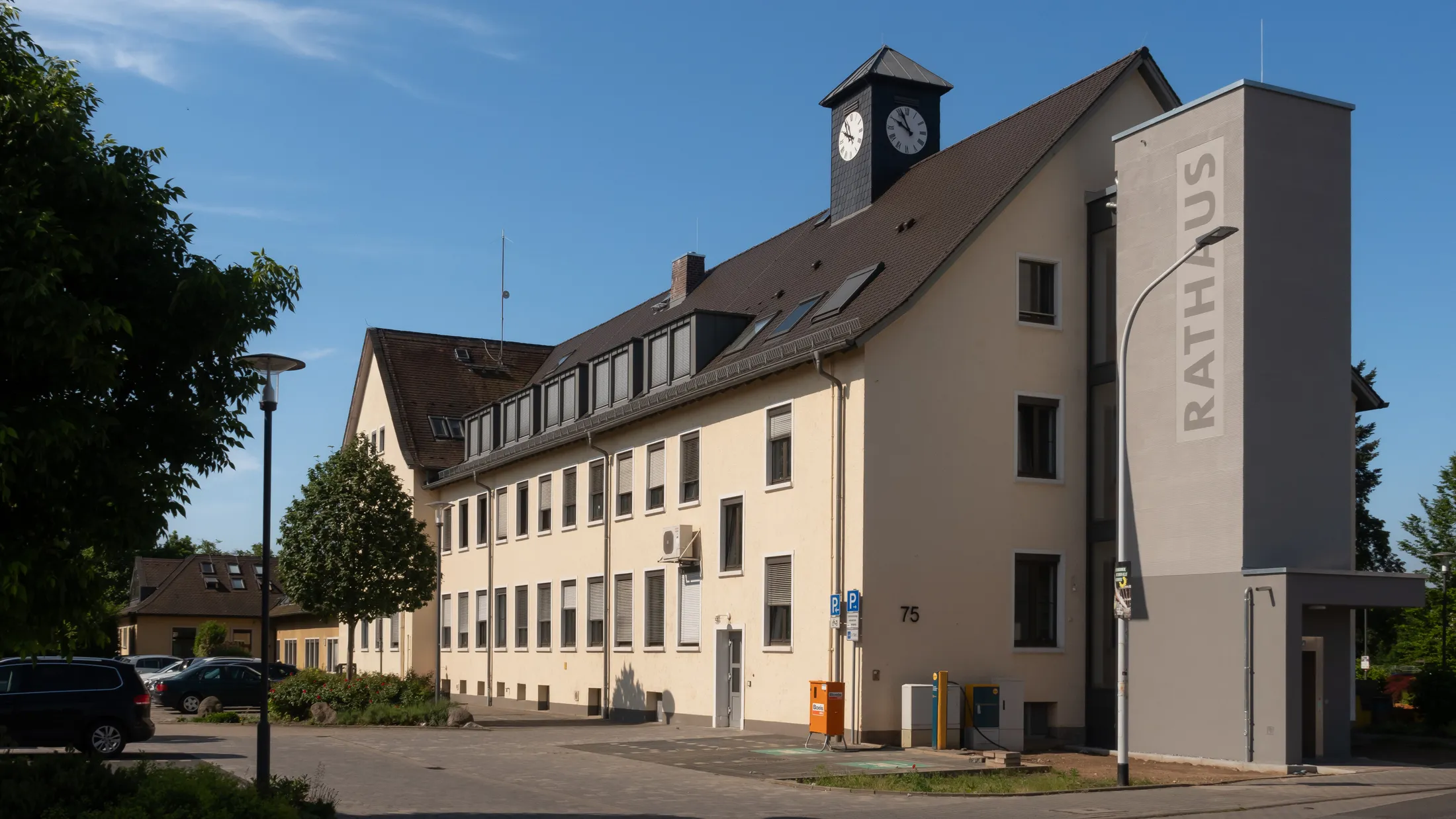 Das Rathaus der Stadt Griesheim in der Wilhelm-Leuschner-Straße 75, ein hellgelber Bau mit Uhrturm auf dem Dach und einem grauen Treppenturm mit der Aufschrift Rathaus, davor ein Parkplatz und Bäume.