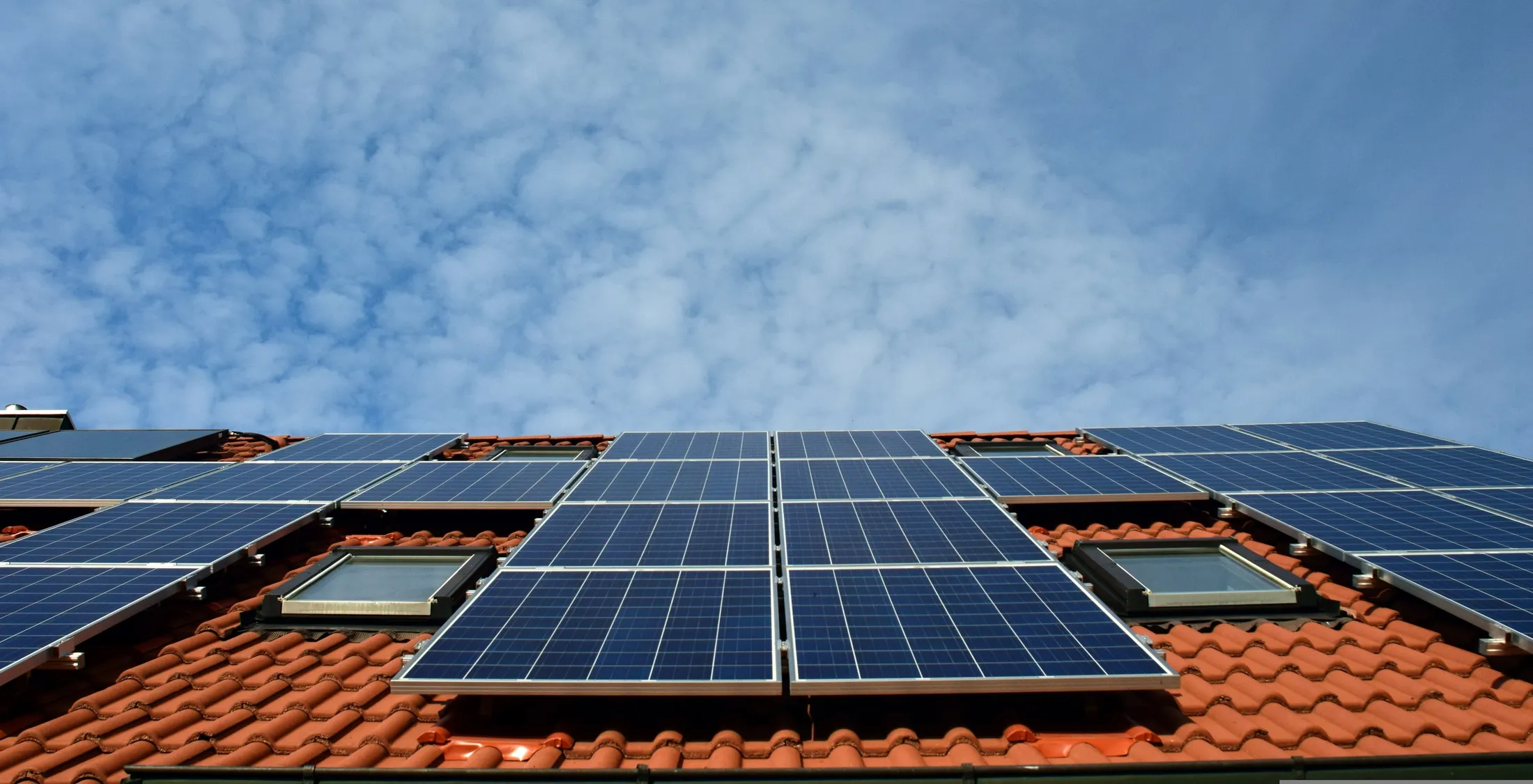 Ziegeldach eines Wohnhauses von unten fotografiert, in zwei Reihen mit Photovoltaikmodulen belegt, dazwischen zwei Dachfenster, darüber blauer Himmel mit Schäfchenwolken.