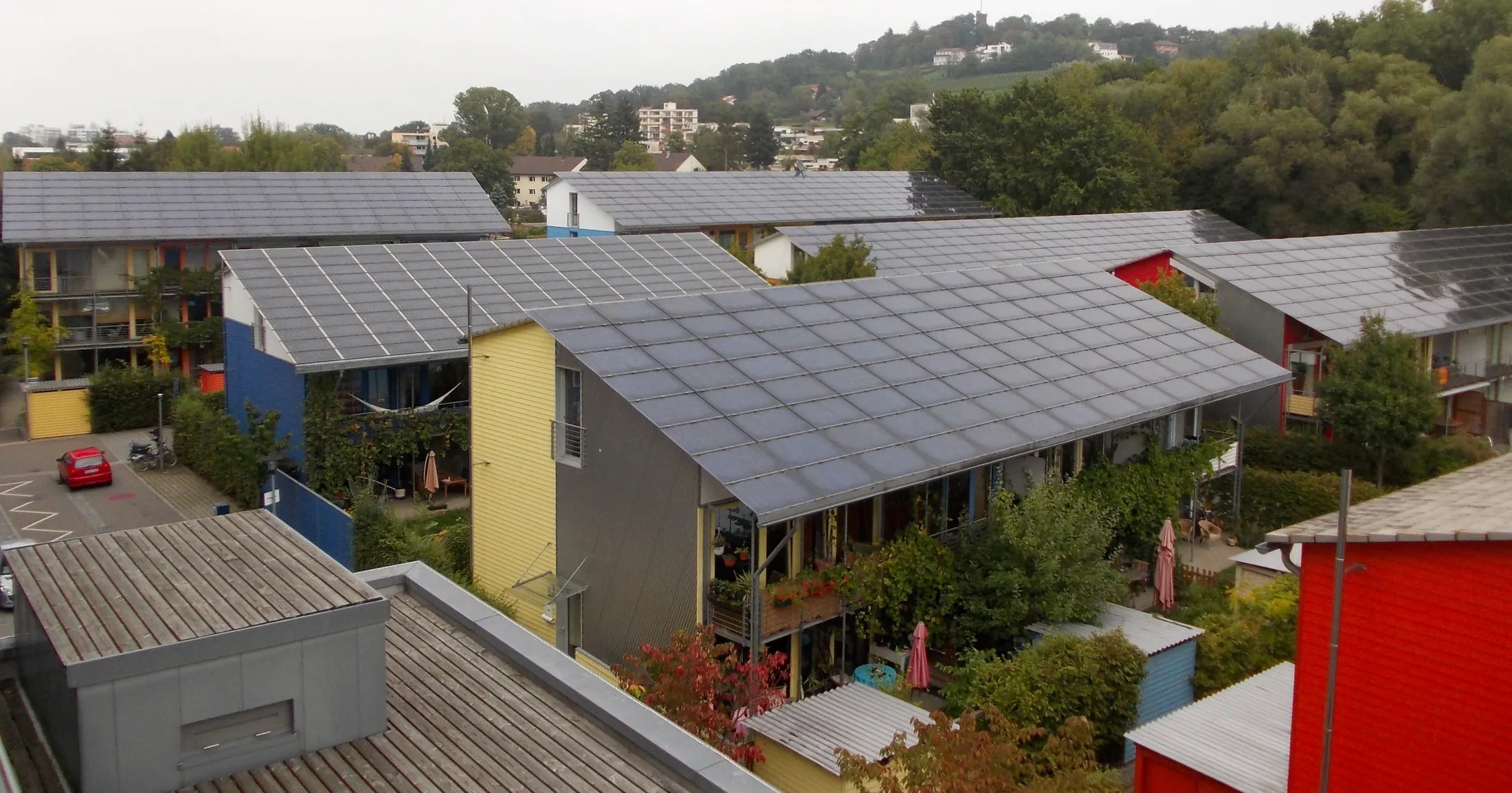 Blick über eine Wohnsiedlung mit bunten Holzfassaden, deren flach geneigte Dächer vollständig mit Photovoltaikmodulen belegt sind, dahinter bewaldete Hügel.